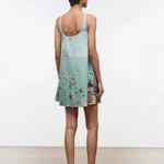 A woman seen from behind wears a light blue Brio Dress, a short tank dress featuring a panoramic beach scene print.