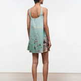 A woman seen from behind wears a light blue Brio Dress, a short tank dress featuring a panoramic beach scene print.