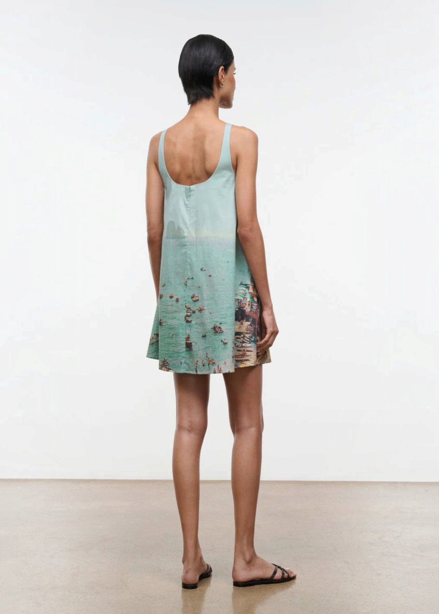 A woman seen from behind wears a light blue Brio Dress, a short tank dress featuring a panoramic beach scene print.