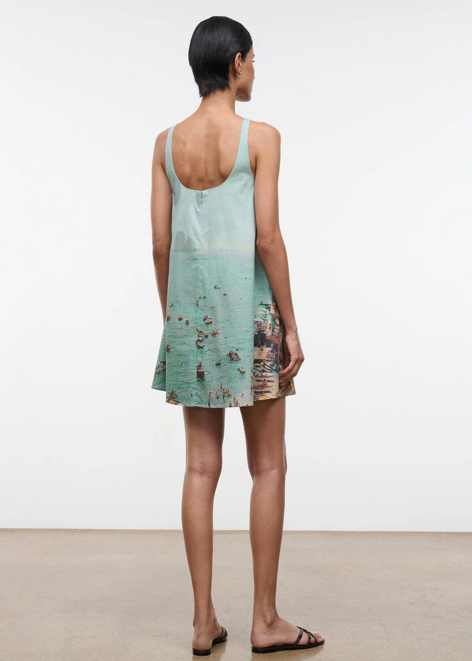 A woman seen from behind wears a light blue Brio Dress, a short tank dress featuring a panoramic beach scene print.
