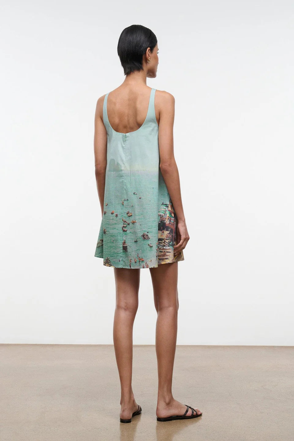 A woman seen from behind wears a light blue Brio Dress, a short tank dress featuring a panoramic beach scene print.