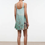 A woman seen from behind wears a light blue Brio Dress, a short tank dress featuring a panoramic beach scene print.