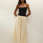 A woman stands against a white textured wall, wearing the Sant Roc Maxi Dress styled as a chic strapless look. She pairs the elegant dress with brown sandals and has long wavy hair, her arms resting behind her back.