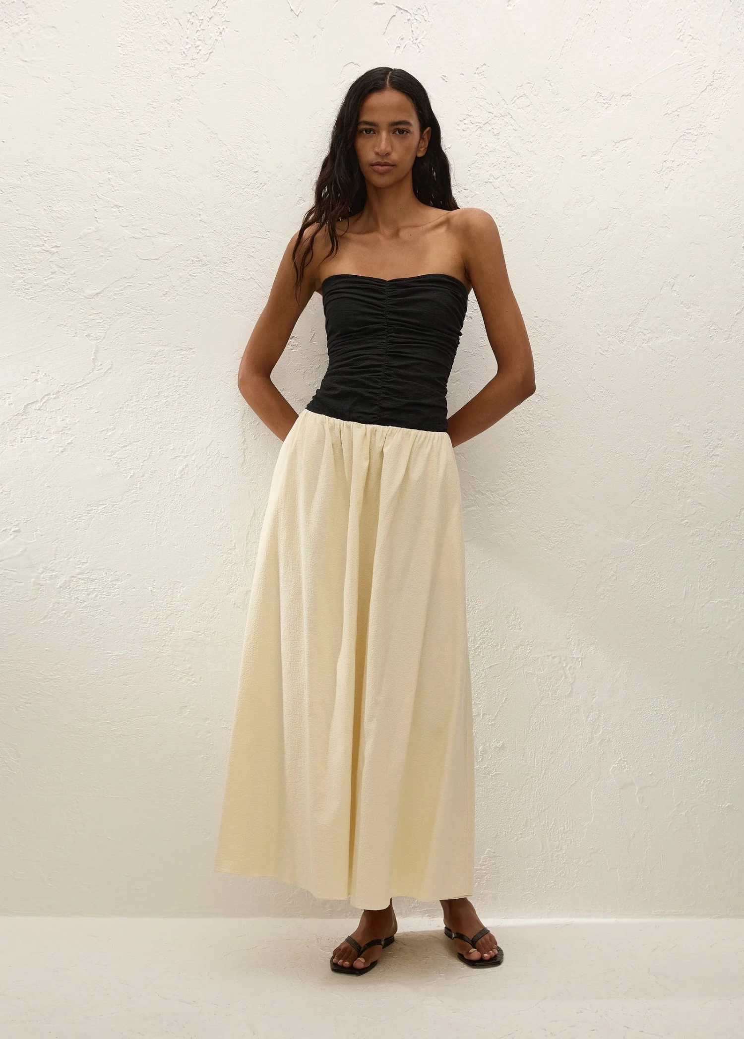 A woman stands against a white textured wall, wearing the Sant Roc Maxi Dress styled as a chic strapless look. She pairs the elegant dress with brown sandals and has long wavy hair, her arms resting behind her back.
