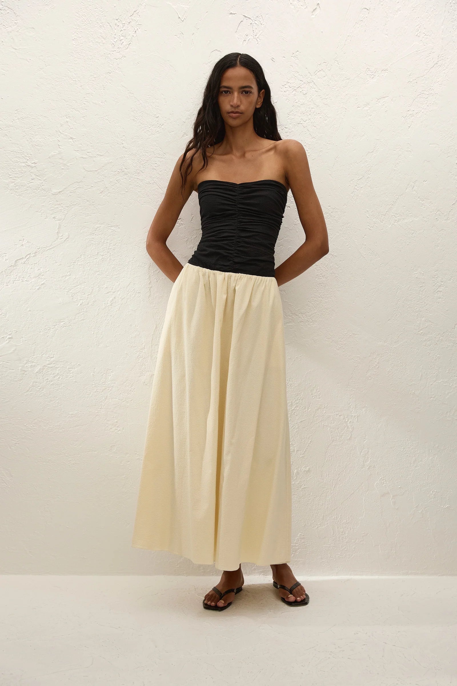 A woman stands against a white textured wall, wearing the Sant Roc Maxi Dress styled as a chic strapless look. She pairs the elegant dress with brown sandals and has long wavy hair, her arms resting behind her back.