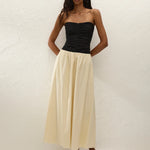 A woman stands against a white textured wall, wearing the Sant Roc Maxi Dress styled as a chic strapless look. She pairs the elegant dress with brown sandals and has long wavy hair, her arms resting behind her back.
