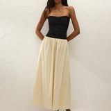 A woman stands against a white textured wall, wearing the Sant Roc Maxi Dress styled as a chic strapless look. She pairs the elegant dress with brown sandals and has long wavy hair, her arms resting behind her back.