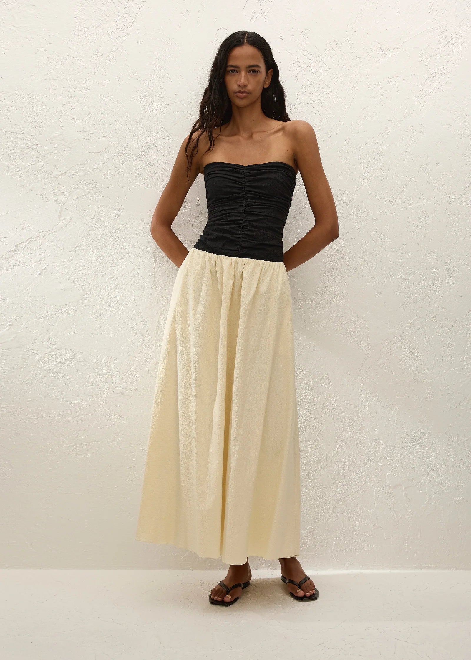 A woman stands against a white textured wall, wearing the Sant Roc Maxi Dress styled as a chic strapless look. She pairs the elegant dress with brown sandals and has long wavy hair, her arms resting behind her back.