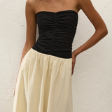 A woman wearing the Sant Roc Maxi Dress—a high-waisted, cream-colored, flowing cotton maxi dress with a strapless black ruched top—stands against a textured white wall.
