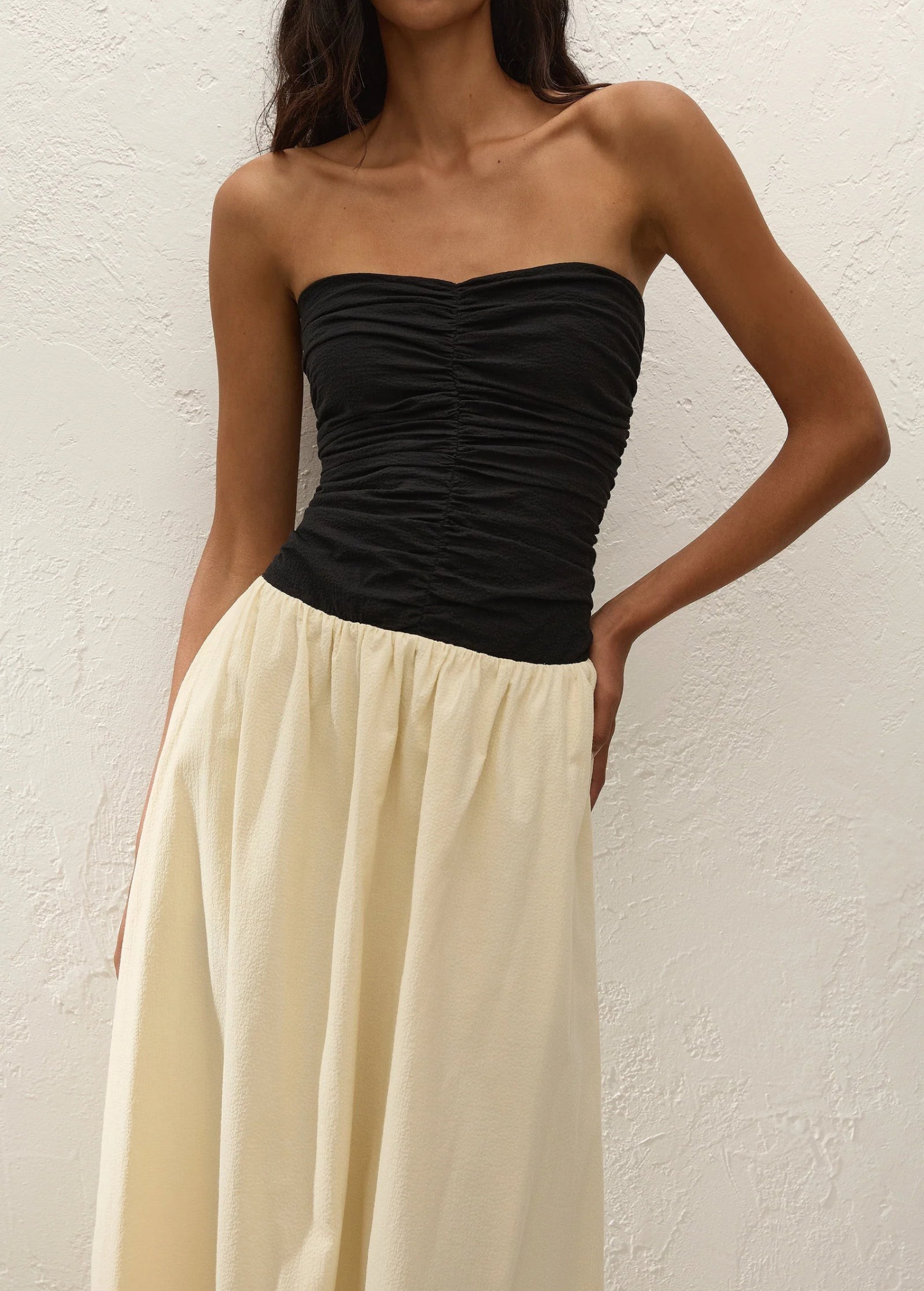 A woman wearing the Sant Roc Maxi Dress—a high-waisted, cream-colored, flowing cotton maxi dress with a strapless black ruched top—stands against a textured white wall.