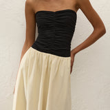 A woman wearing the Sant Roc Maxi Dress—a high-waisted, cream-colored, flowing cotton maxi dress with a strapless black ruched top—stands against a textured white wall.