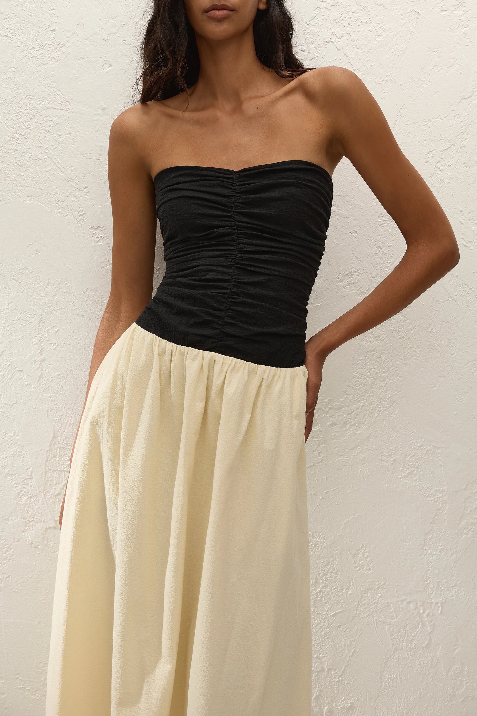 A woman wearing the Sant Roc Maxi Dress—a high-waisted, cream-colored, flowing cotton maxi dress with a strapless black ruched top—stands against a textured white wall.