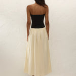 A woman stands facing a textured white wall, wearing the Sant Roc Maxi Dress—a black strapless dress with a long cream skirt—paired with black sandals. She has dark, wavy hair and her arms are relaxed by her sides.