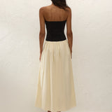 A woman stands facing a textured white wall, wearing the Sant Roc Maxi Dress—a black strapless dress with a long cream skirt—paired with black sandals. She has dark, wavy hair and her arms are relaxed by her sides.