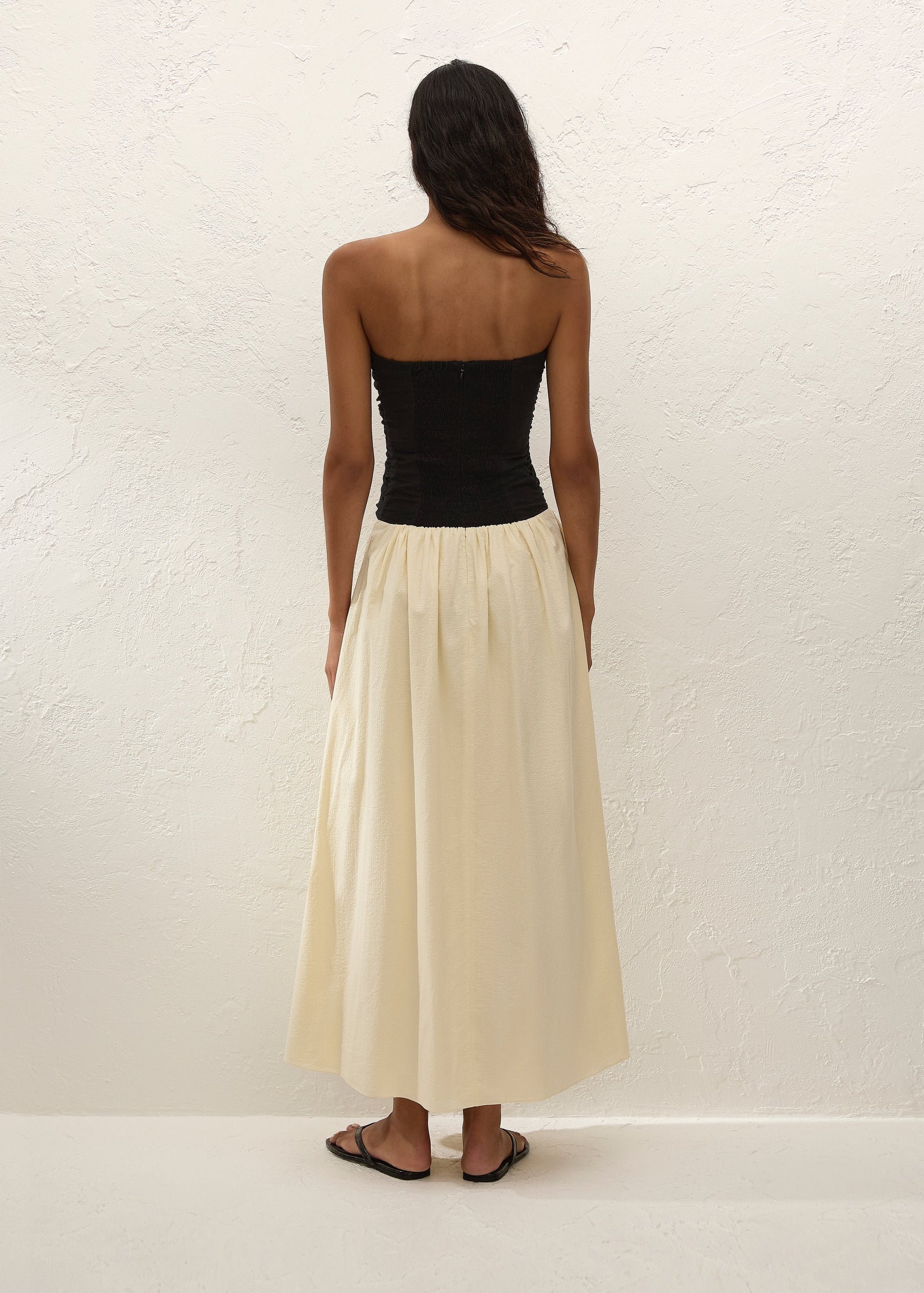 A woman stands facing a textured white wall, wearing the Sant Roc Maxi Dress—a black strapless dress with a long cream skirt—paired with black sandals. She has dark, wavy hair and her arms are relaxed by her sides.
