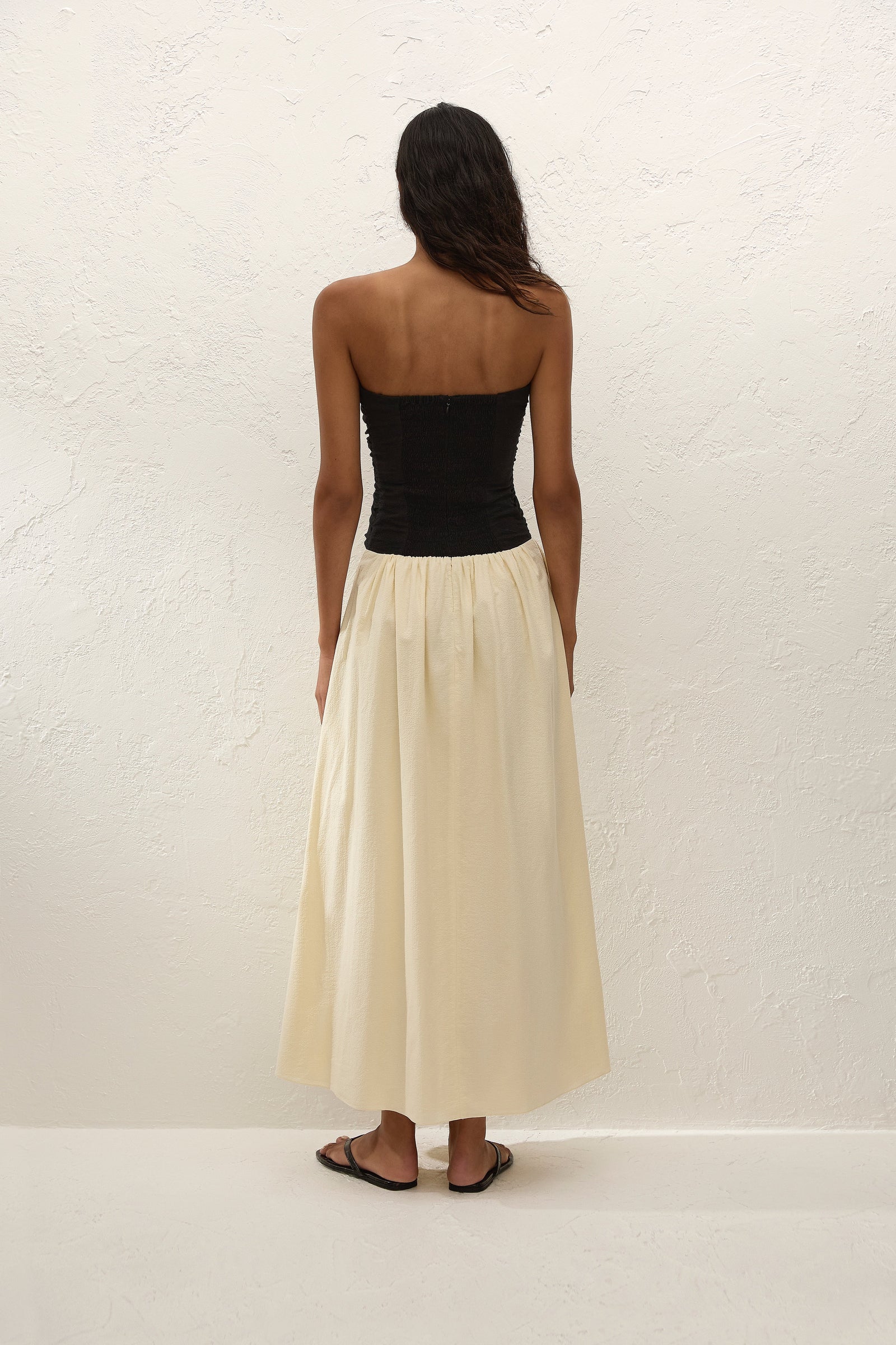 A woman stands facing a textured white wall, wearing the Sant Roc Maxi Dress—a black strapless dress with a long cream skirt—paired with black sandals. She has dark, wavy hair and her arms are relaxed by her sides.