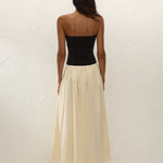 A woman stands facing a textured white wall, wearing the Sant Roc Maxi Dress—a black strapless dress with a long cream skirt—paired with black sandals. She has dark, wavy hair and her arms are relaxed by her sides.