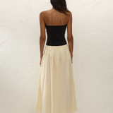 A woman stands facing a textured white wall, wearing the Sant Roc Maxi Dress—a black strapless dress with a long cream skirt—paired with black sandals. She has dark, wavy hair and her arms are relaxed by her sides.