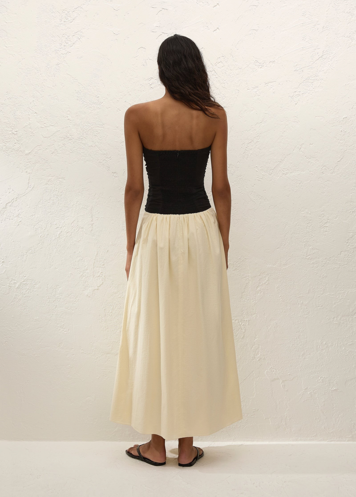 A woman stands facing a textured white wall, wearing the Sant Roc Maxi Dress—a black strapless dress with a long cream skirt—paired with black sandals. She has dark, wavy hair and her arms are relaxed by her sides.