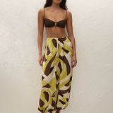 A woman wears a brown bikini top paired with Kortez Balloon Pants, which have a relaxed balloon silhouette and an abstract geometric pattern in shades of green, brown, and cream.