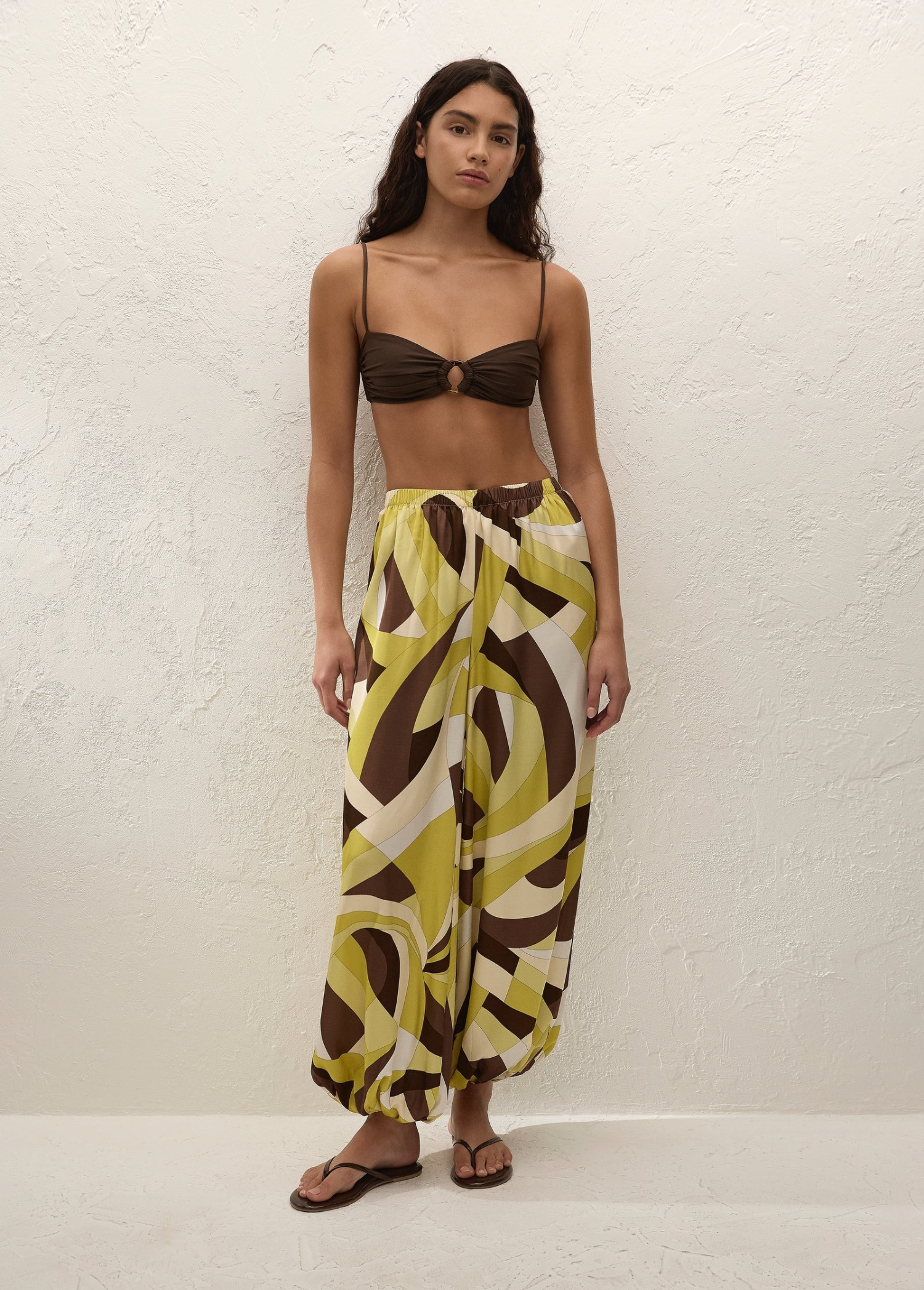 A woman wears a brown bikini top paired with Kortez Balloon Pants, which have a relaxed balloon silhouette and an abstract geometric pattern in shades of green, brown, and cream.