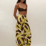 A person wears high-waisted, balloon-leg Kortez Balloon Pants with a relaxed fit and a geometric pattern in green, white, and brown, paired with a brown bikini top and thong sandals.