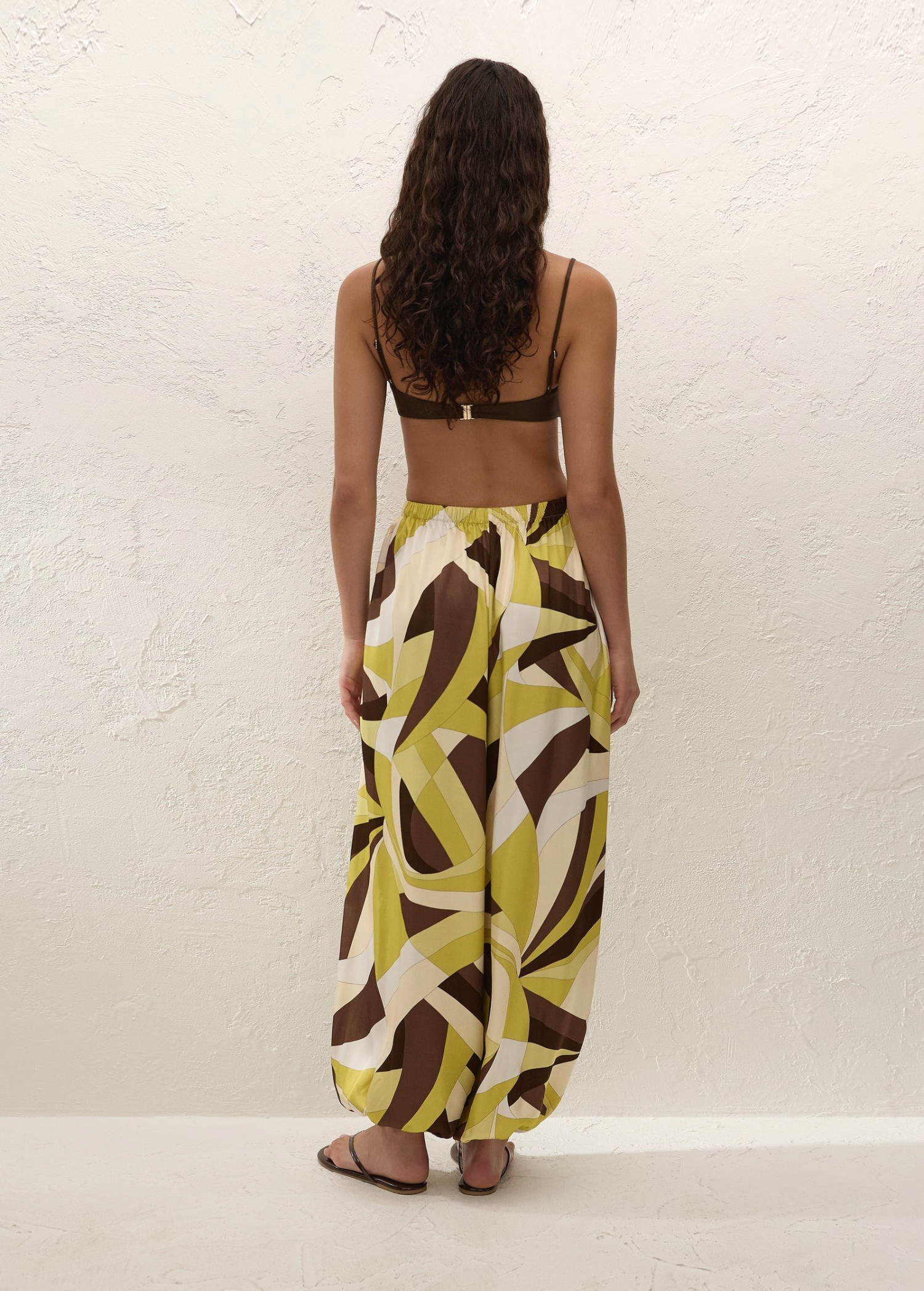 A person viewed from behind wearing a brown bikini top and Kortez Balloon Pants, which feature a relaxed, high-waisted fit and a green, white, and brown geometric pattern.
