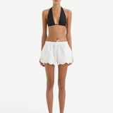 A person wearing a black bikini top and white Klara Mini Short drawstring shorts with a scalloped hem and floral cut-out embroidery.