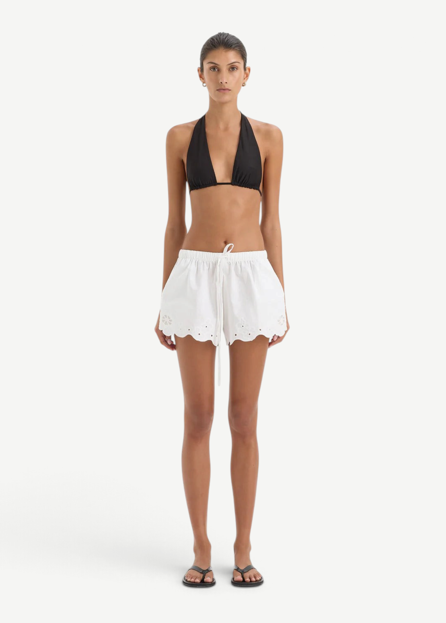 A person wearing a black bikini top and white Klara Mini Short drawstring shorts with a scalloped hem and floral cut-out embroidery.