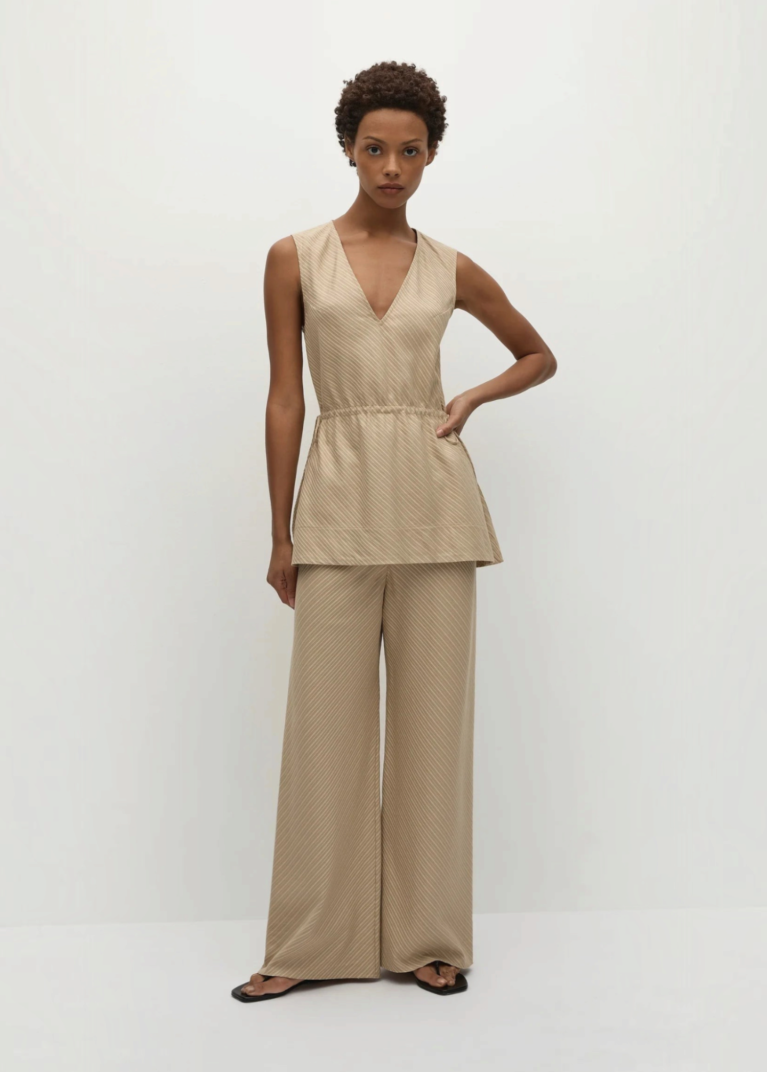 A person wearing a beige pinstripe Selan Top with a v-neck and matching wide-leg trousers, creating a relaxed silhouette with a refined tunic.