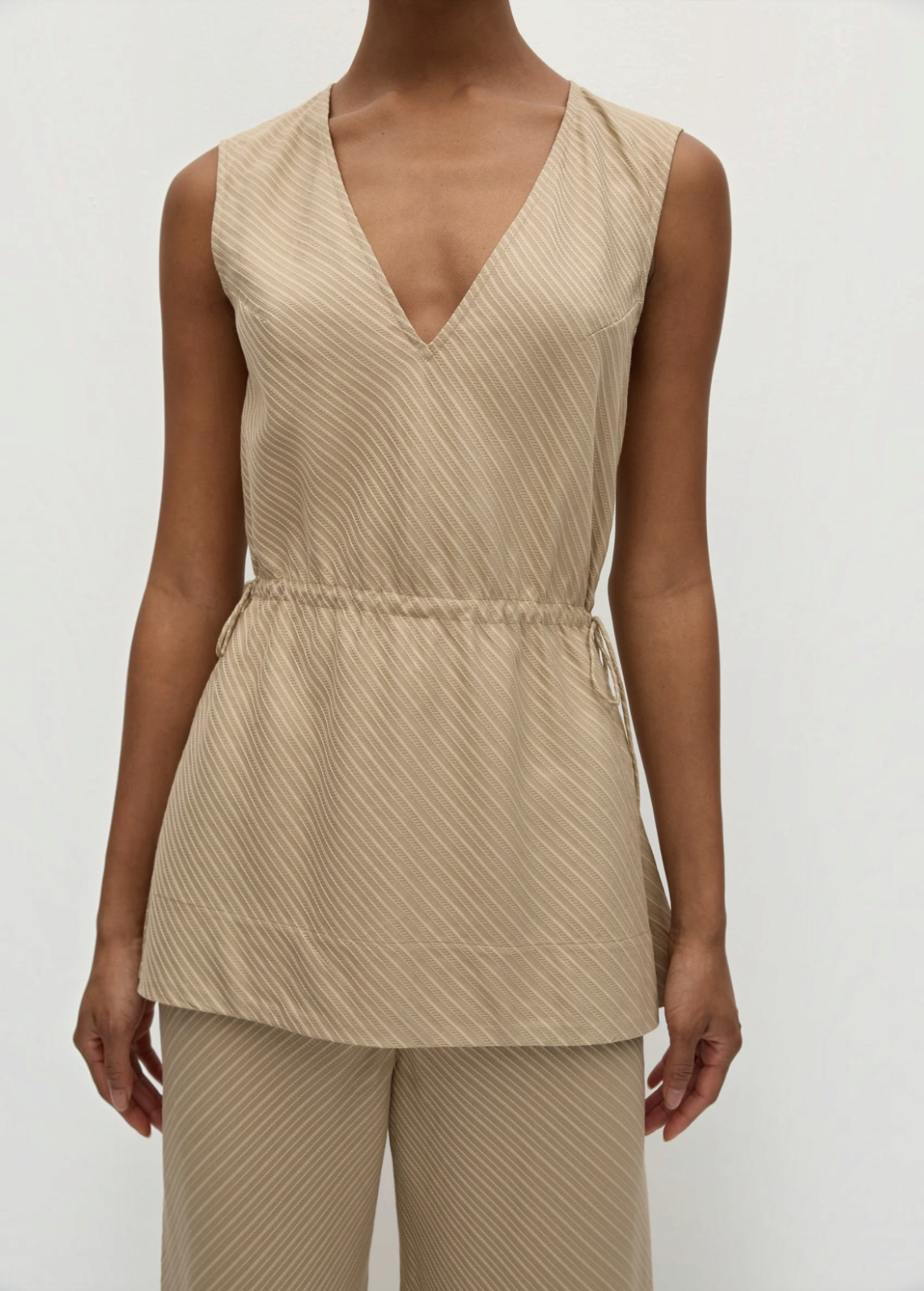 Selan Top in beige, featuring a sleeveless V-neck, relaxed silhouette, drawstring waist, and a diagonal textured pinstripe pattern.