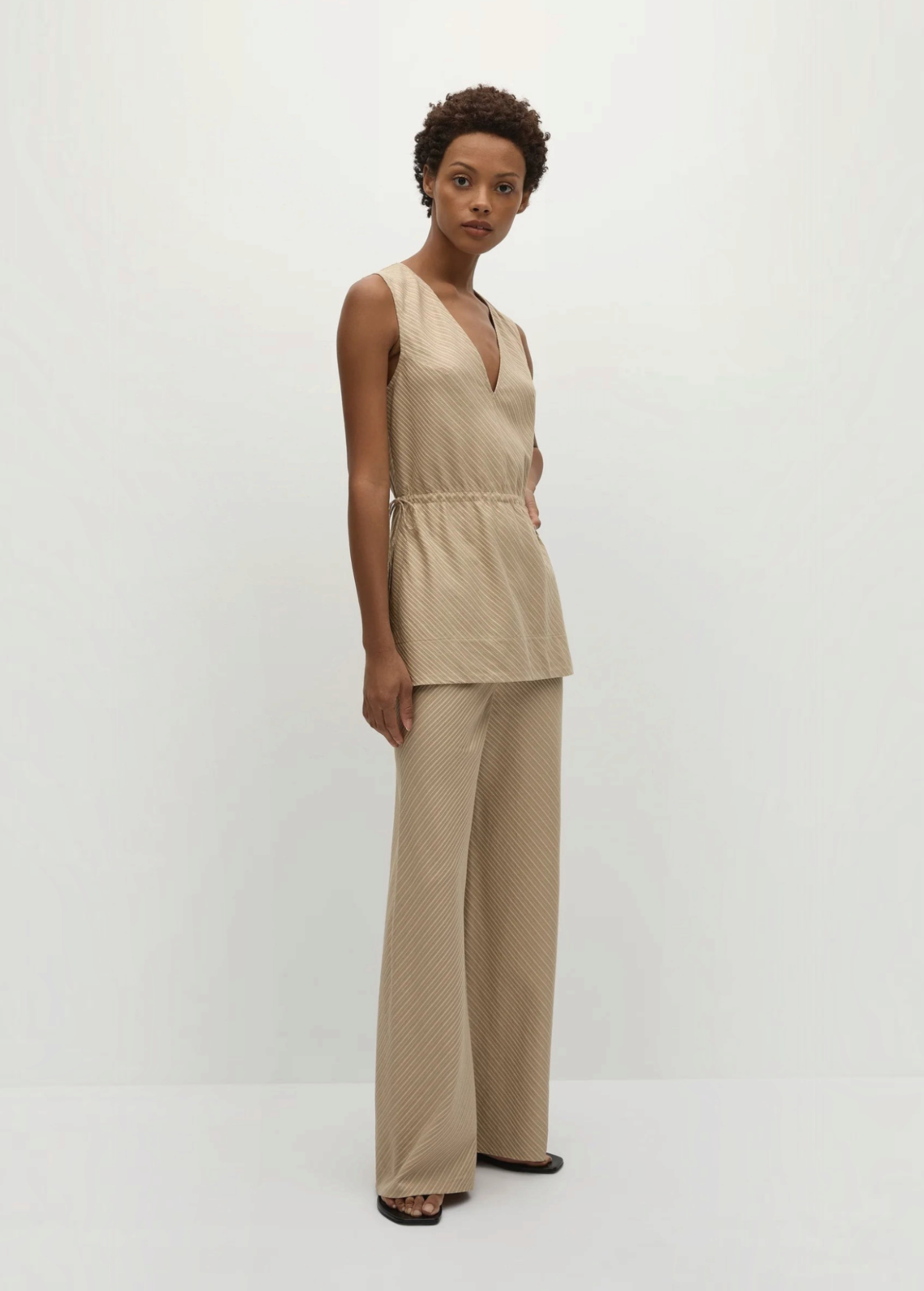 A woman wears a beige Selan Top, a sleeveless V-neck tunic with a drawstring waist, paired with matching pinstripe wide-leg trousers.