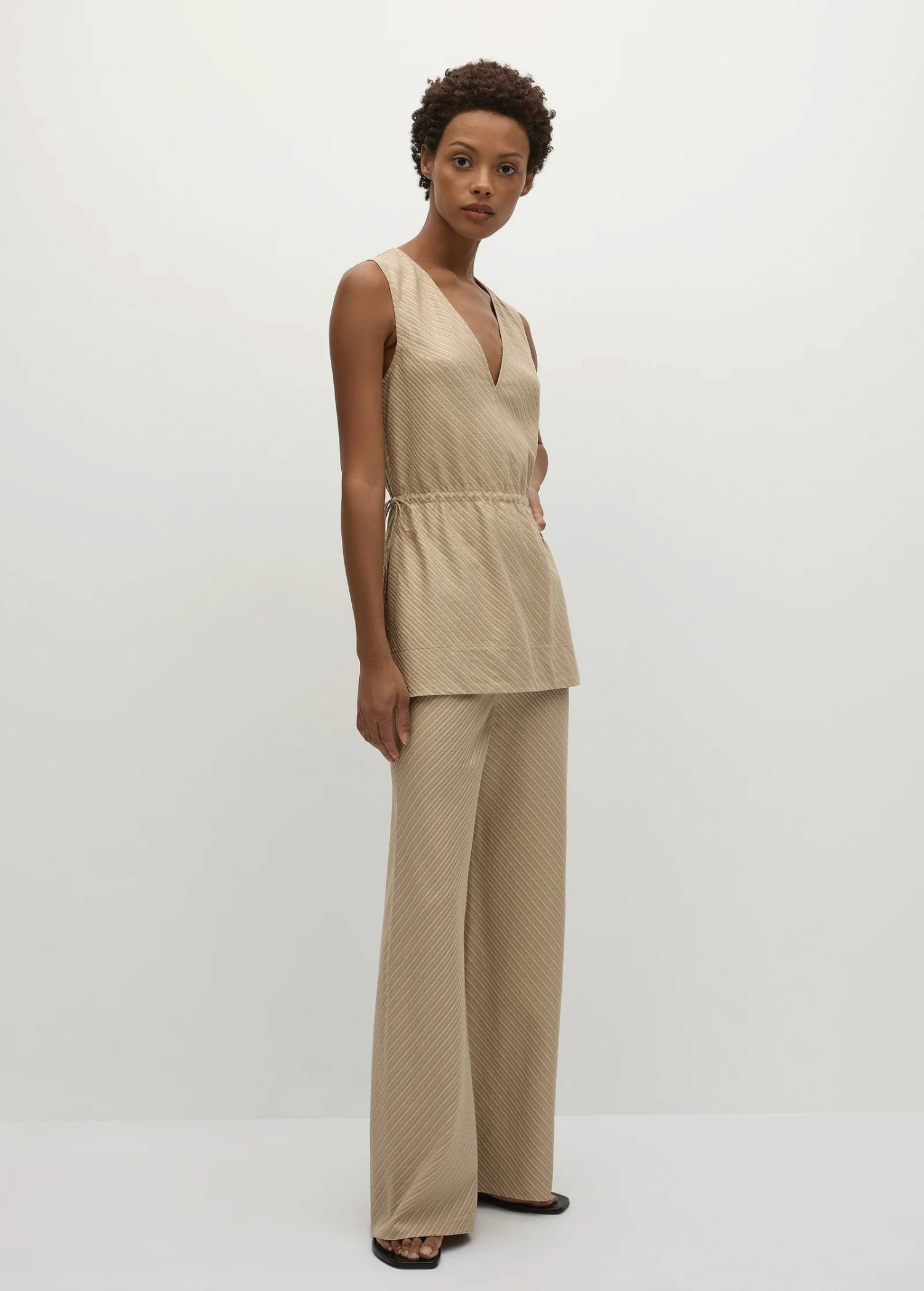A woman wears a beige Selan Top, a sleeveless V-neck tunic with a drawstring waist, paired with matching pinstripe wide-leg trousers.