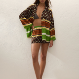 A person wearing a Cora Shirt and matching shorts in a brown, green, and cream geometric pattern over a patterned bikini top, paired with flip-flops.