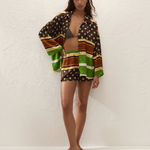 A person wearing a Cora Shirt and matching shorts in a brown, green, and cream geometric pattern over a patterned bikini top, paired with flip-flops.