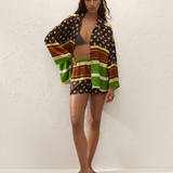 A person wearing a Cora Shirt and matching shorts in a brown, green, and cream geometric pattern over a patterned bikini top, paired with flip-flops.
