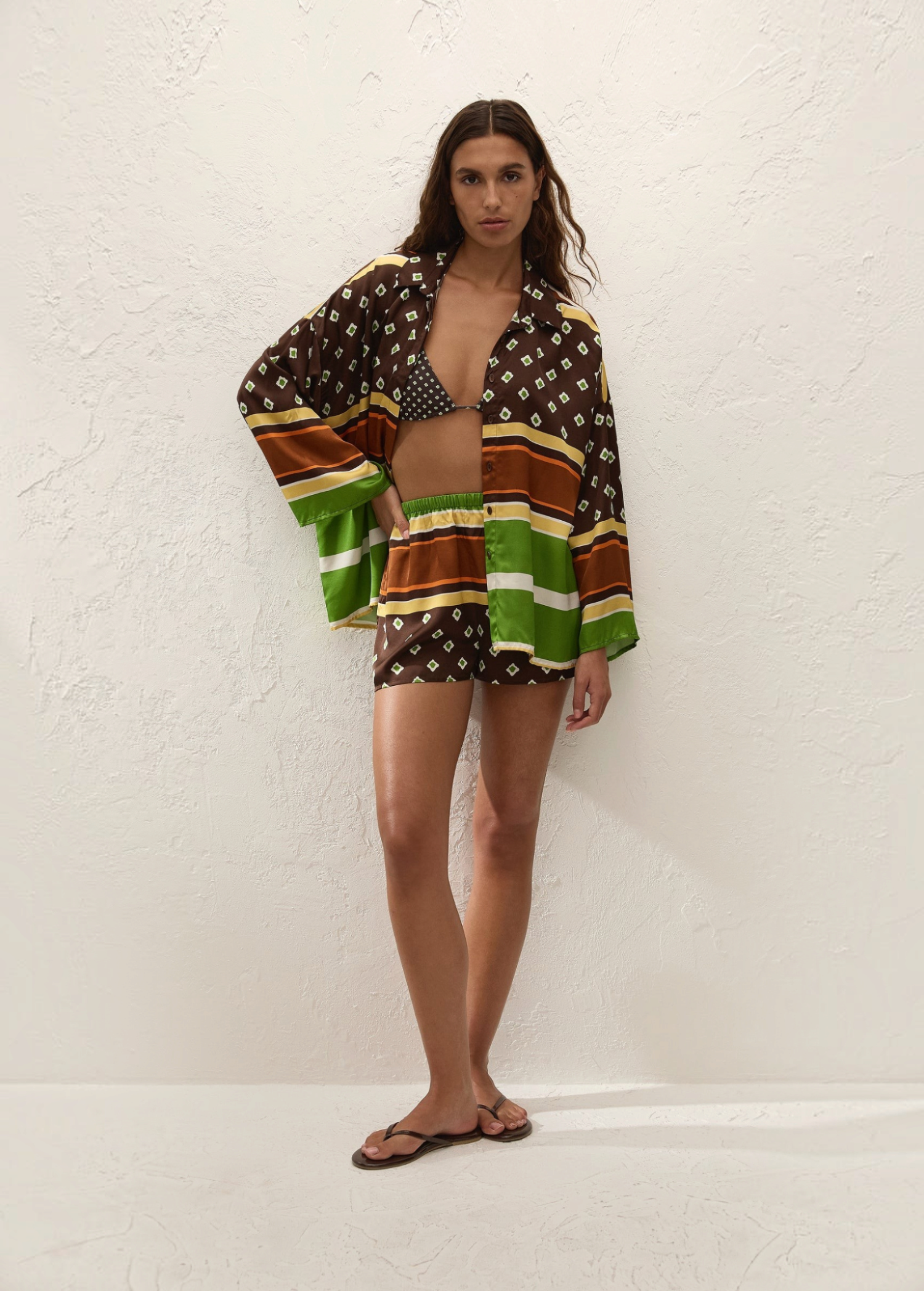 A person wearing a Cora Shirt and matching shorts in a brown, green, and cream geometric pattern over a patterned bikini top, paired with flip-flops.