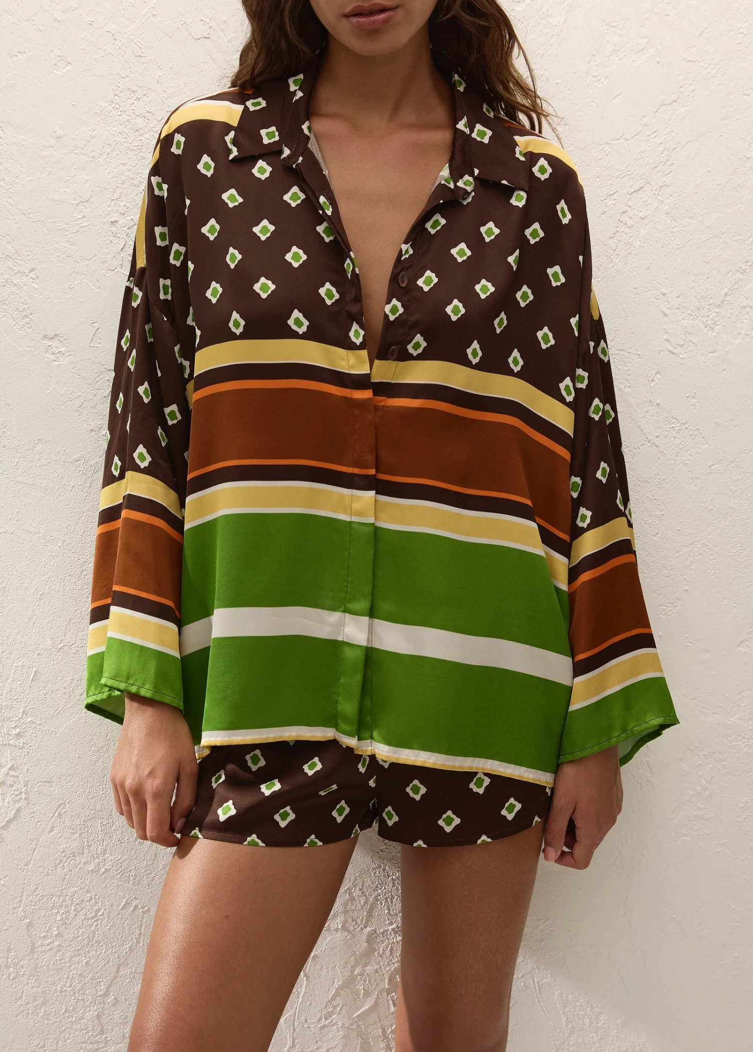 A person wearing a loose-fitting Cora Shirt and matching shorts featuring a brown and green geometric pattern with horizontal stripes.