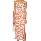 Woman wearing the Chloe One Shoulder Midi Dress, featuring a white, one-shoulder design with orange and red floral prints. The dress is crafted from lightweight linen and fully lined for comfort. She holds a small handbag and wears sandals, standing facing forward against a plain white background.