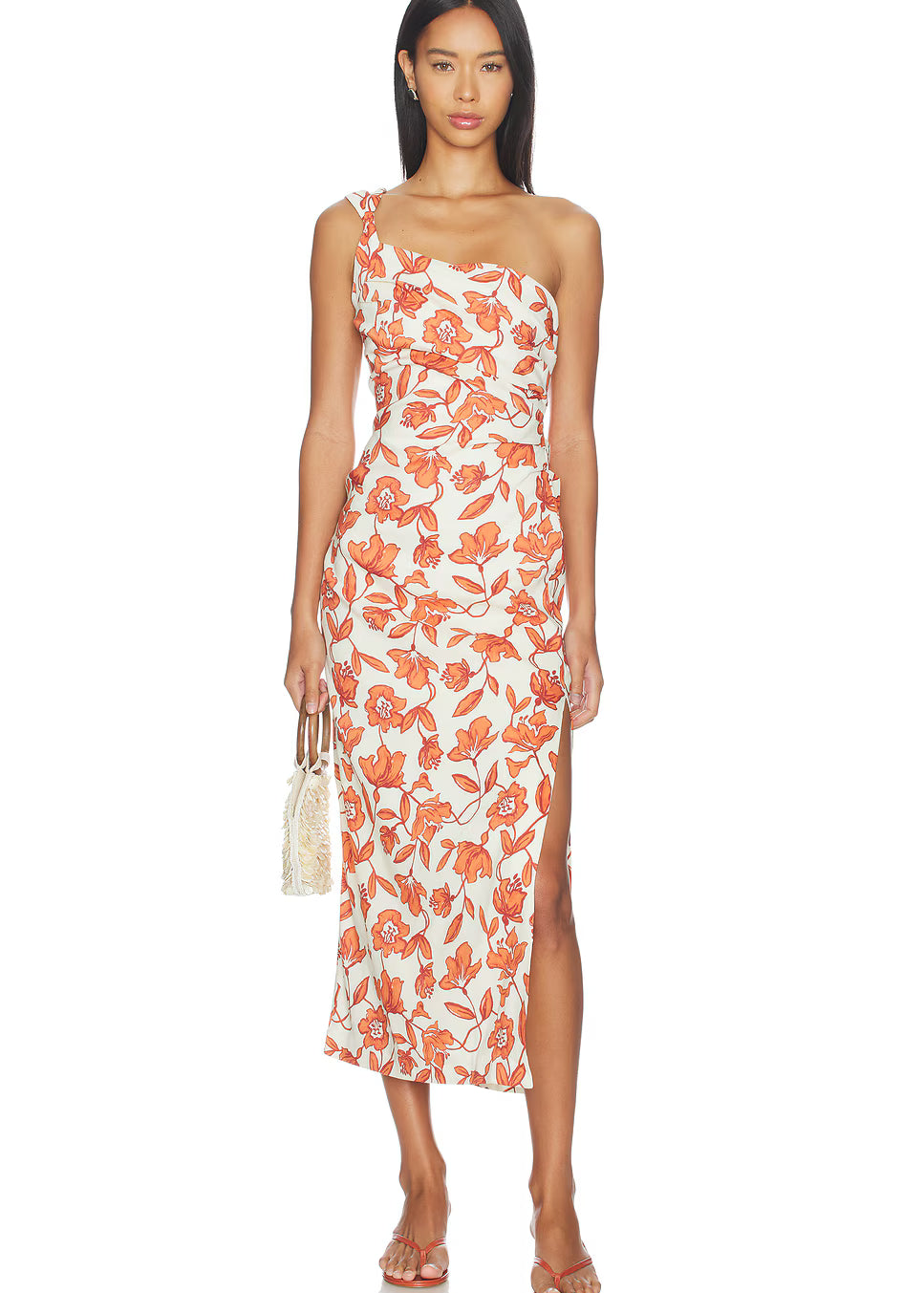 A woman models the Chloe One Shoulder Midi Dress—a sleeveless, one-shoulder white midi dress featuring a bold orange floral print, side slit, and crafted from lightweight linen fabric made in Turkey. The fully lined dress is styled with flat sandals and a small woven handbag as she stands against a white background.