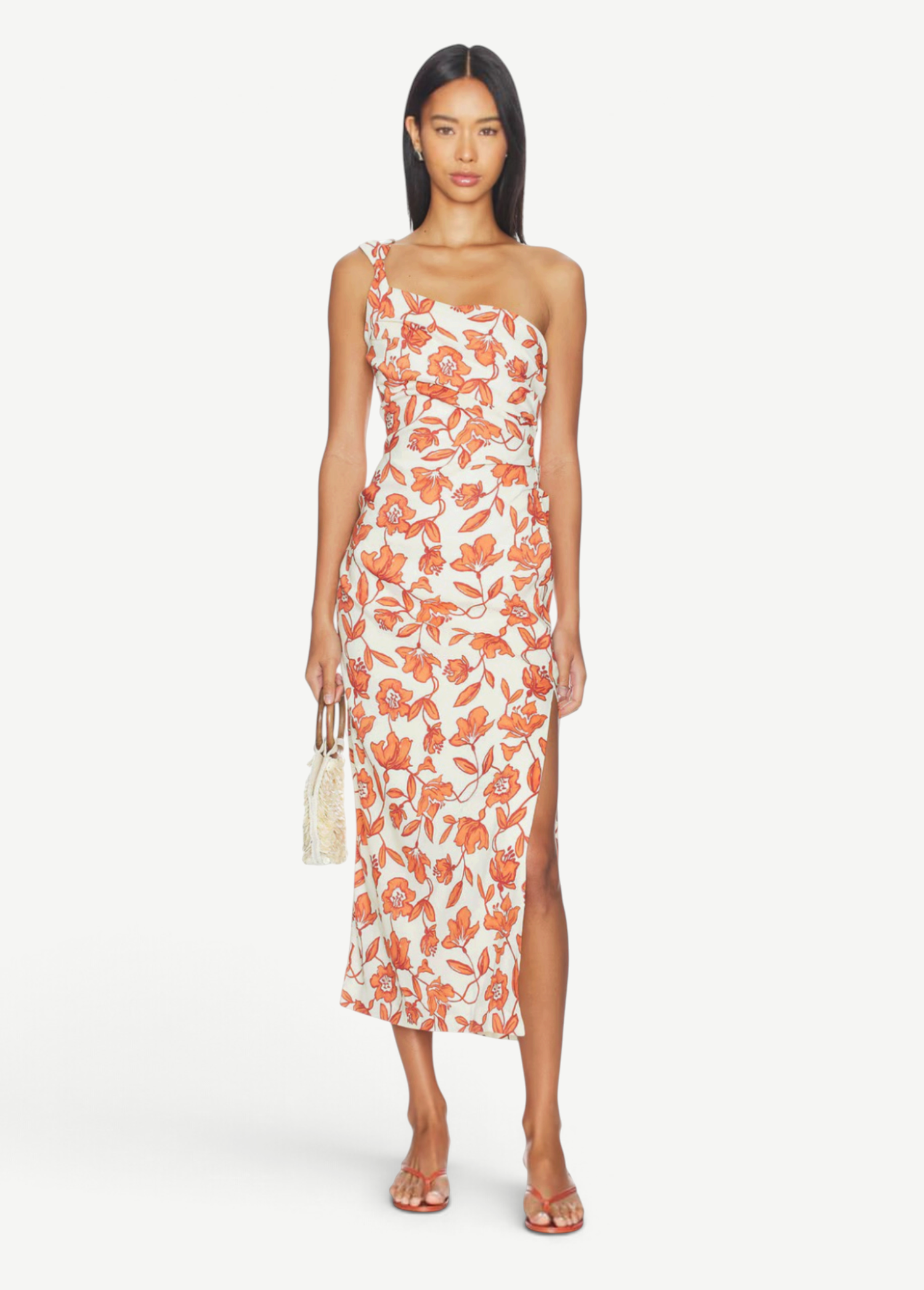 A woman models the Chloe One Shoulder Midi Dress—a sleeveless, one-shoulder white midi dress featuring a bold orange floral print, side slit, and crafted from lightweight linen fabric made in Turkey. The fully lined dress is styled with flat sandals and a small woven handbag as she stands against a white background.