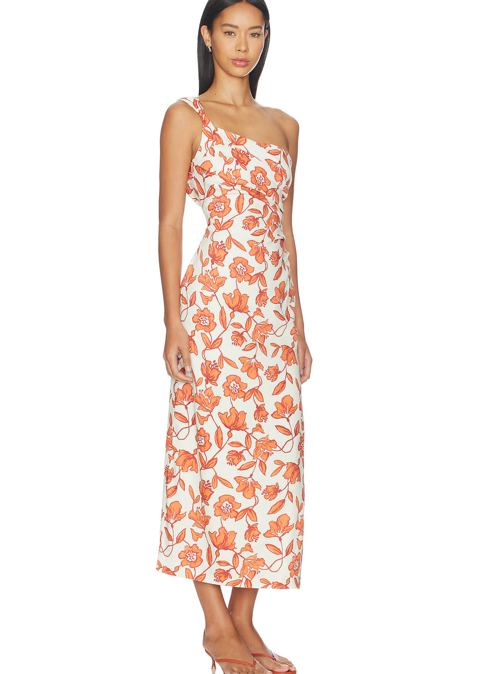 A woman models the Chloe One Shoulder Midi Dress—an ankle-length, white dress featuring an orange floral pattern, crafted from lightweight linen and fully lined for comfort. She pairs it with flat sandals and stands against a plain white background.