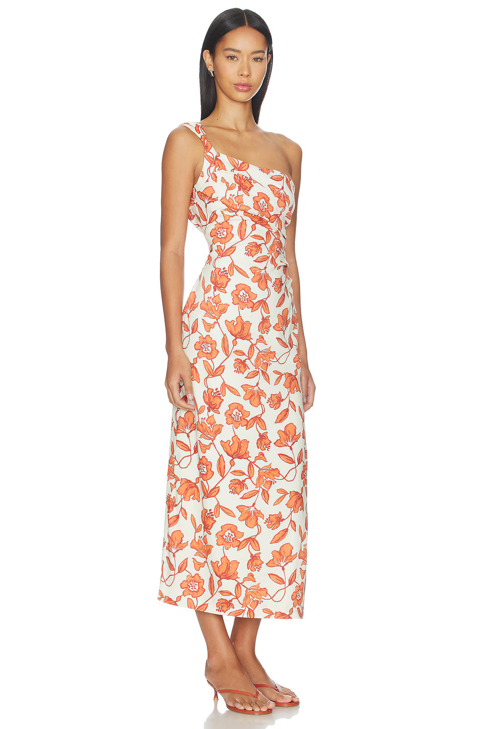 A woman models the Chloe One Shoulder Midi Dress—an ankle-length, white dress featuring an orange floral pattern, crafted from lightweight linen and fully lined for comfort. She pairs it with flat sandals and stands against a plain white background.