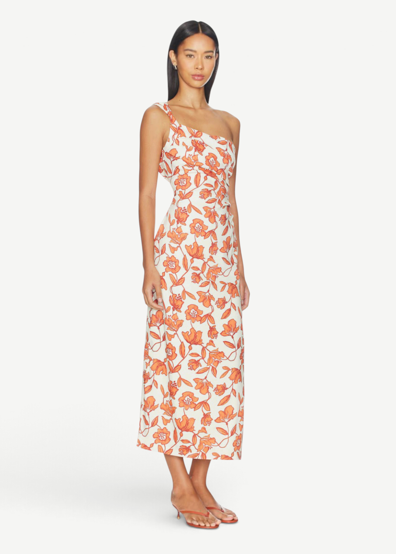 A woman models the Chloe One Shoulder Midi Dress—an ankle-length, white dress featuring an orange floral pattern, crafted from lightweight linen and fully lined for comfort. She pairs it with flat sandals and stands against a plain white background.