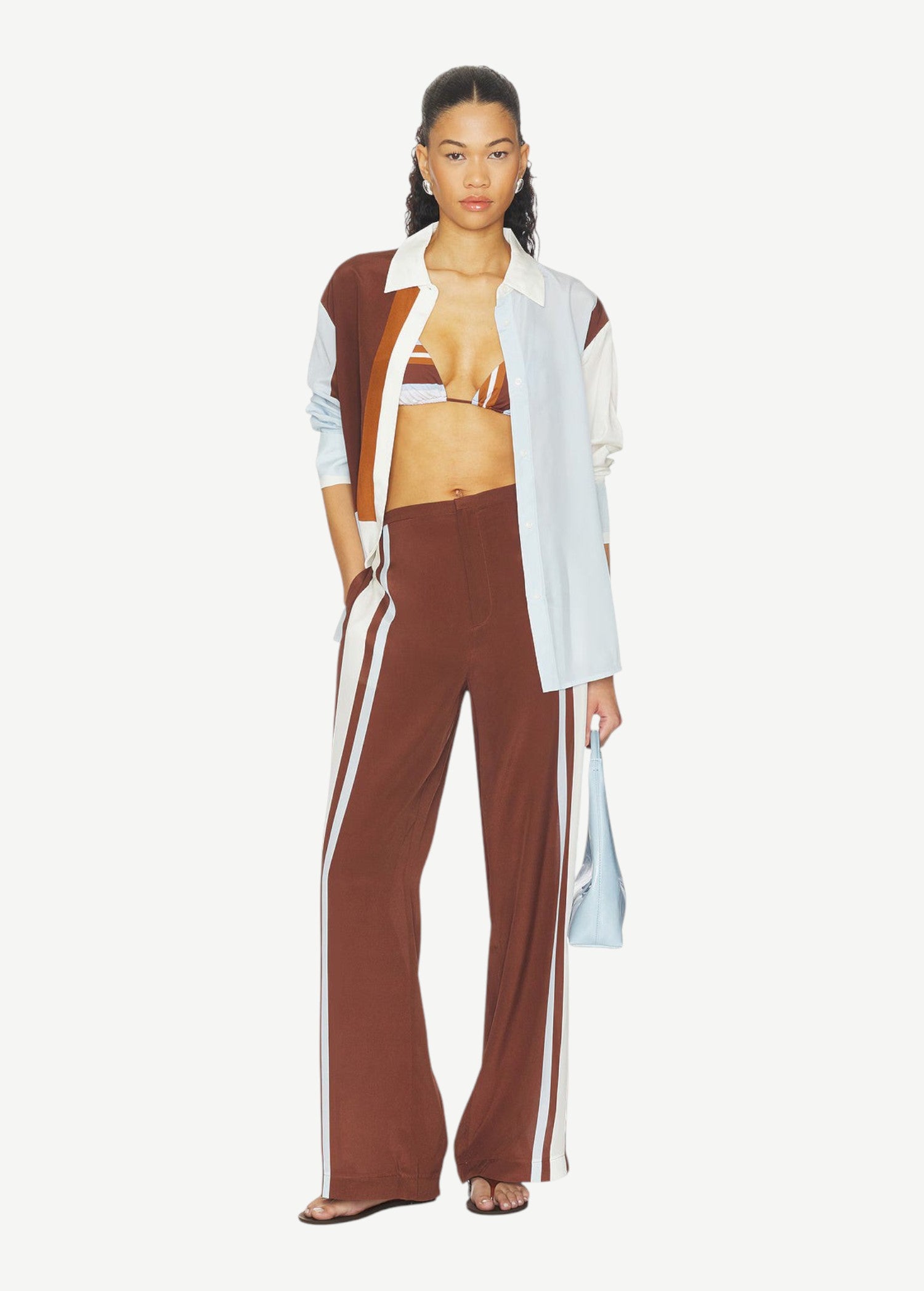A person wearing Marais Trouser wide-leg silk trousers with white side stripes, an unbuttoned color-blocked shirt, and a striped bikini top.