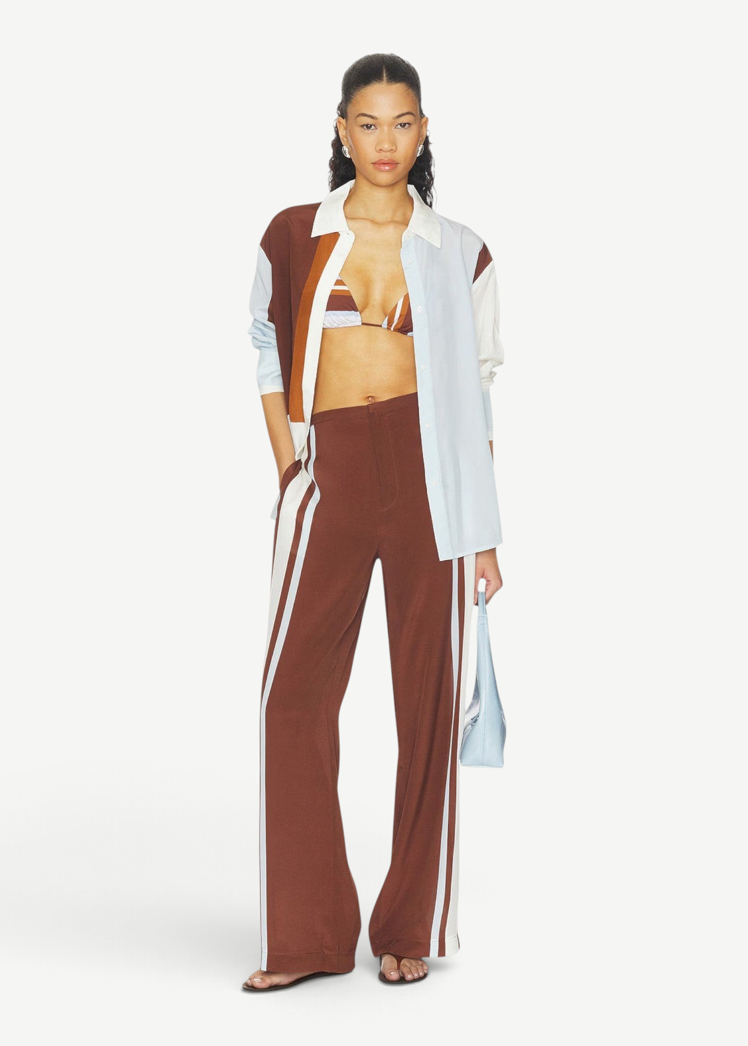 A person wearing Marais Trouser wide-leg silk trousers with white side stripes, an unbuttoned color-blocked shirt, and a striped bikini top.