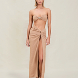 A woman stands facing forward with her arms at her sides and a neutral expression, wearing a tan bandeau top and the Gwen Skirt—a matching high-slit skirt-sarong—paired with sandals, set against a plain beige background.