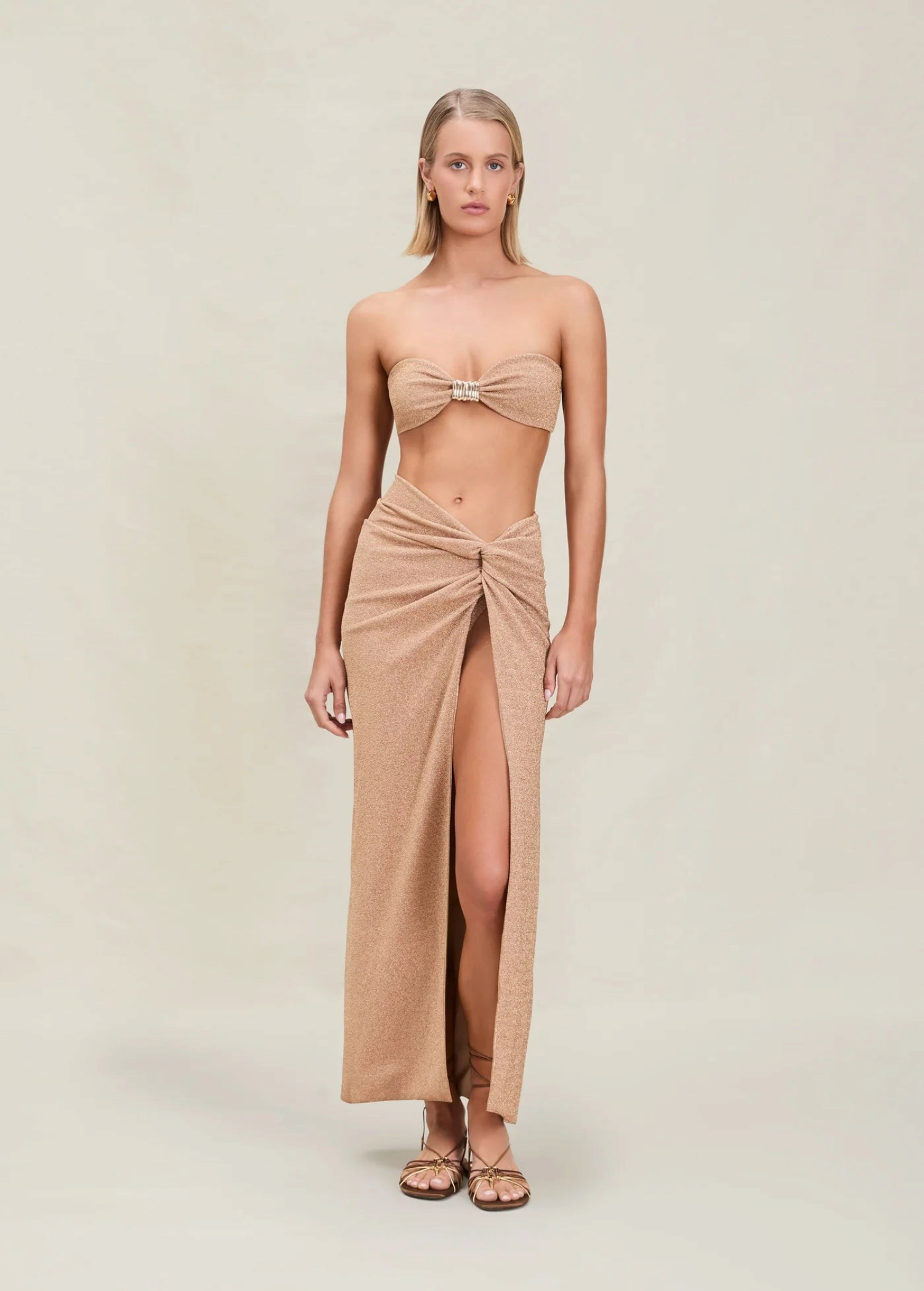 A woman stands facing forward with her arms at her sides and a neutral expression, wearing a tan bandeau top and the Gwen Skirt—a matching high-slit skirt-sarong—paired with sandals, set against a plain beige background.