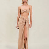 A woman stands facing forward with her arms at her sides and a neutral expression, wearing a tan bandeau top and the Gwen Skirt—a matching high-slit skirt-sarong—paired with sandals, set against a plain beige background.