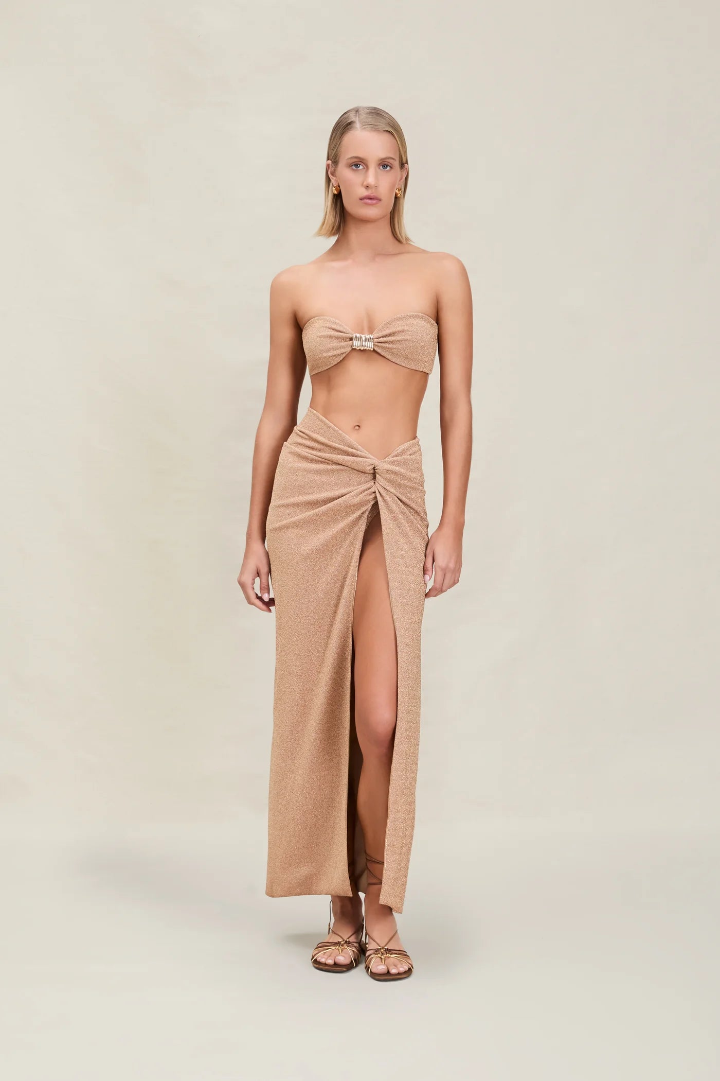 A woman stands facing forward with her arms at her sides and a neutral expression, wearing a tan bandeau top and the Gwen Skirt—a matching high-slit skirt-sarong—paired with sandals, set against a plain beige background.